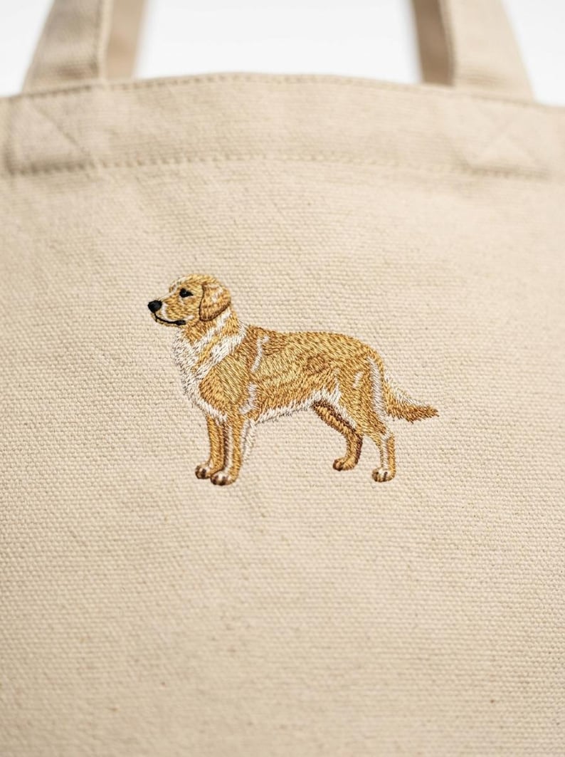 May include: Beige tote bag featuring an embroidered golden retriever dog. The dog is depicted in a standing pose with detailed stitching in shades of gold, brown, and white. The bag has a simple design with sturdy handles.