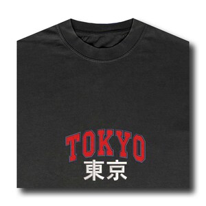 Puede incluir: Una camiseta gris oscuro con la palabra "TOKYO" en rojo, contorneada en azul, y los caracteres japoneses de Tokio en blanco. La camiseta tiene cuello redondo y est&aacute; doblada.