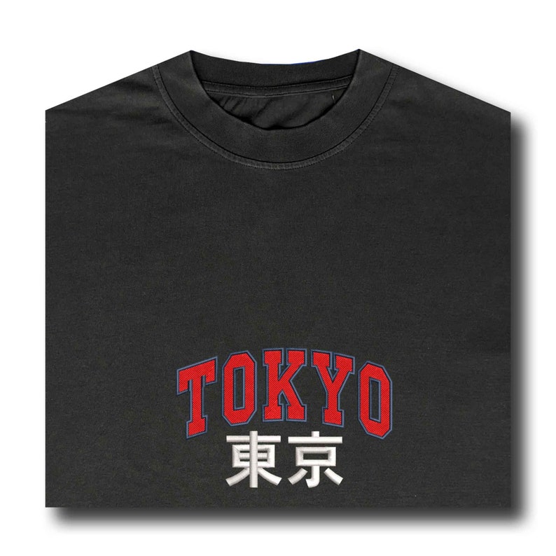 Puede incluir: Una camiseta gris oscuro con la palabra "TOKYO" en rojo, contorneada en azul, y los caracteres japoneses de Tokio en blanco. La camiseta tiene cuello redondo y est&aacute; doblada.