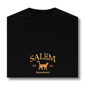 K&ouml;nnte beinhalten: Schwarzes T-Shirt mit einem goldenen gestickten Logo, auf dem "SALEM EST. 1626 Massachusetts" steht und eine Silhouette einer Katze abgebildet ist.