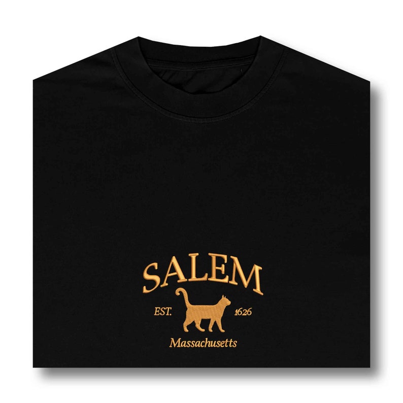K&ouml;nnte beinhalten: Schwarzes T-Shirt mit einem goldenen gestickten Logo, auf dem "SALEM EST. 1626 Massachusetts" steht und eine Silhouette einer Katze abgebildet ist.