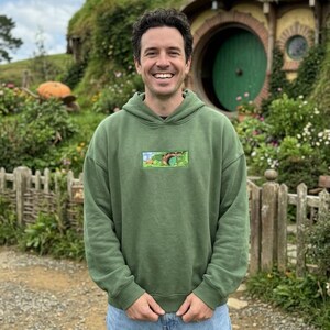 May include: A person wearing a green hoodie with a small rectangular graphic. The hoodie is paired with blue jeans. The background features a green hillside with a round green door and a wooden fence.