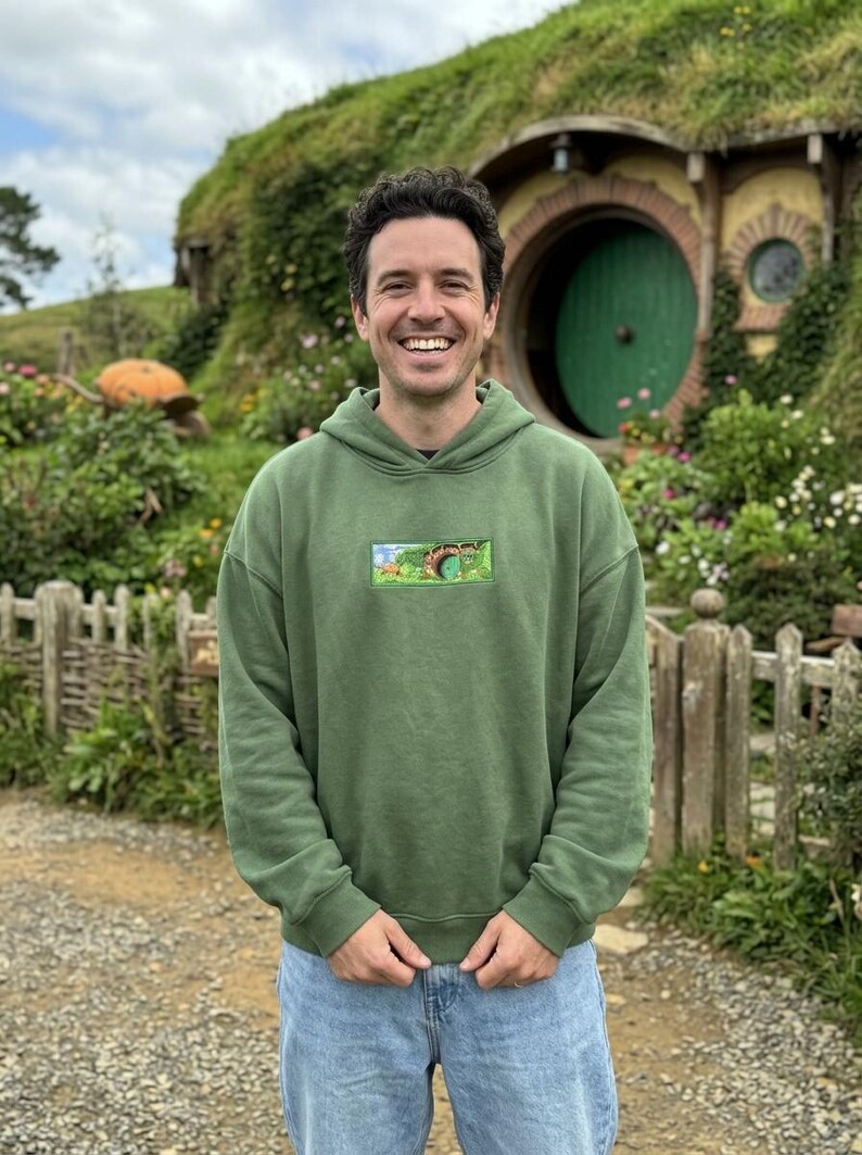 May include: A person wearing a green hoodie with a small rectangular graphic. The hoodie is paired with blue jeans. The background features a green hillside with a round green door and a wooden fence.