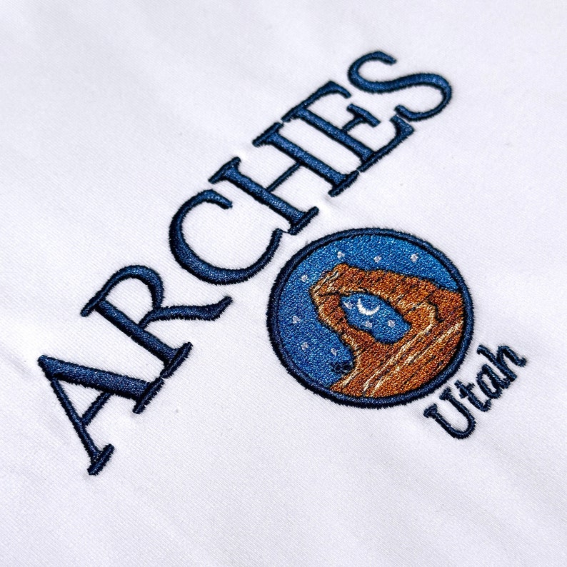 Pode incluir: Um tecido branco com texto bordado em azul escuro que diz ARCHES e Utah. Entre as palavras, h&aacute; um emblema circular que retrata uma forma&ccedil;&atilde;o de arco de rocha marrom contra um c&eacute;u noturno azul cheio de estrelas brancas e uma lua crescente.