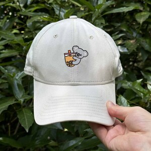 May include: A white baseball cap with a grey koala holding a cup of boba tea. The koala is embroidered on the front of the cap.