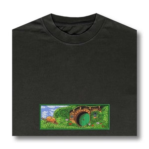 May include: Dark gray t-shirt with a rectangular embroidered patch. The patch depicts a green door, a wheelbarrow, and a grassy landscape with a blue sky. The design is inspired by a hobbit hole.