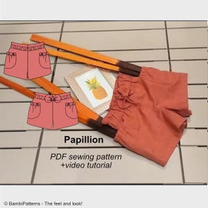 Può includere: Un'illustrazione digitale di un cartamodello per un paio di pantaloncini chiamato "Papillion". I pantaloncini sono di colore pesca e hanno un fiocco in vita. Il testo "Papillion" e "Cartamodello PDF + tutorial video" è incluso anche nell'immagine.