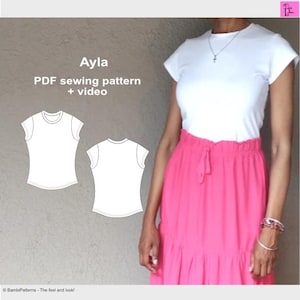 May include: A PDF sewing pattern for a simple t-shirt called "Ayla" with a white background and black lines. The pattern includes a video tutorial. The image also shows a woman wearing a white t-shirt and a pink skirt.