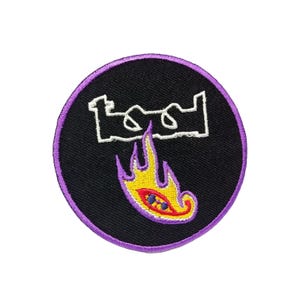 以下が含まれることがあります： 紫色の縁取りが付いた丸い黒いパッチ。パッチには、赤と青の目のデザインの黄色い炎の上に「TOOL」という単語の白いアウトラインが表示されています。パッチはおそらく衣類やアクセサリー用です。