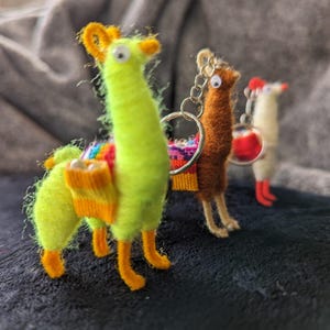 Puede incluir: Tres llaveros de llama hechos a mano. El primero es verde lima con detalles en naranja y una manta colorida. El segundo es marrón con un llavero. El tercero es blanco con detalles en rojo.