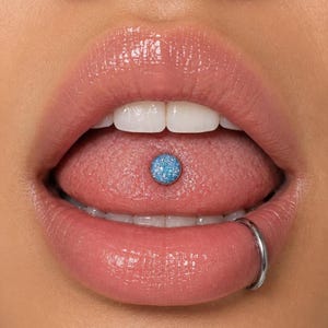 May include: A tongue piercing featuring a circular, light blue, glittery stud set in a metallic base, positioned centrally on the tongue. Additionally, a smooth, thin, silver-colored metal hoop ring is inserted through the lower lip.