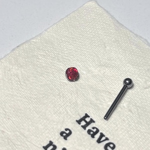 May include: A body jewelry piece consisting of a small, round, red iridescent stone setting and a separate silver-toned metal post with a smooth, spherical end. The items are positioned on a textured white surface with partial black text visible.