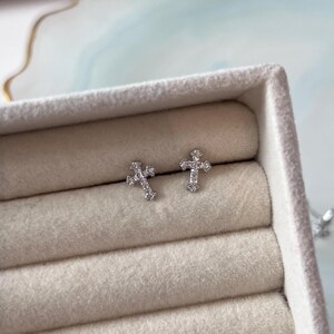 Peut inclure: Une paire de boucles d'oreilles à clous en forme de croix, chacune ornée de petites pierres transparentes et scintillantes incrustées sur la surface du motif. Les boucles d'oreilles sont placées côte à côte, révélant une finition métallique.