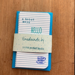 May include: A set of small notebooks with blue and white striped pages, secured by a light blue band. The notebooks feature the words "A Quick Note" and "Hello". The band is printed with "Handmade by secret pocket books" and a teacup illustration.