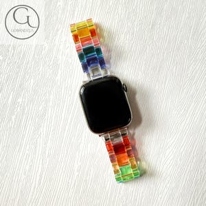 May include: A smartwatch with a black rectangular screen and a multi-colored, translucent resin link band. The band features a vibrant gradient of red, orange, yellow, green, blue, and purple segments. The logo GOBANDZUS is visible in the top left corner.