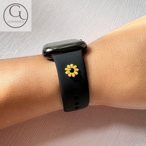 Op de afbeelding: Een zwarte Apple Watch met een zwarte band en een gele bloembedel. Het horloge zit om de pols van een persoon.