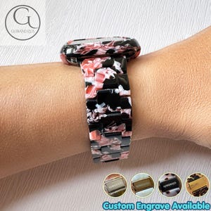 Könnte beinhalten: Ein Uhrenarmband mit einem marmorierten Muster aus schwarzen, weißen und rosafarbenen Harzgliedern. Das Band verfügt über einen rechteckigen Anschluss und einen sicheren Verschluss. Vier kleine Kreissymbole unten zeigen Metallverschlüsse.