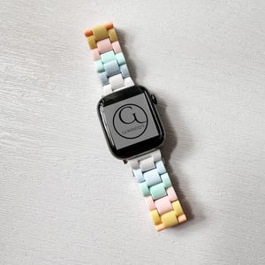 Op de afbeelding: Een witte Apple Watch met een regenboogkleurig siliconen bandje. Het horloge heeft een zwart wijzerplaat met de letter "G" en de tekst "GOMANDZUS" eronder.