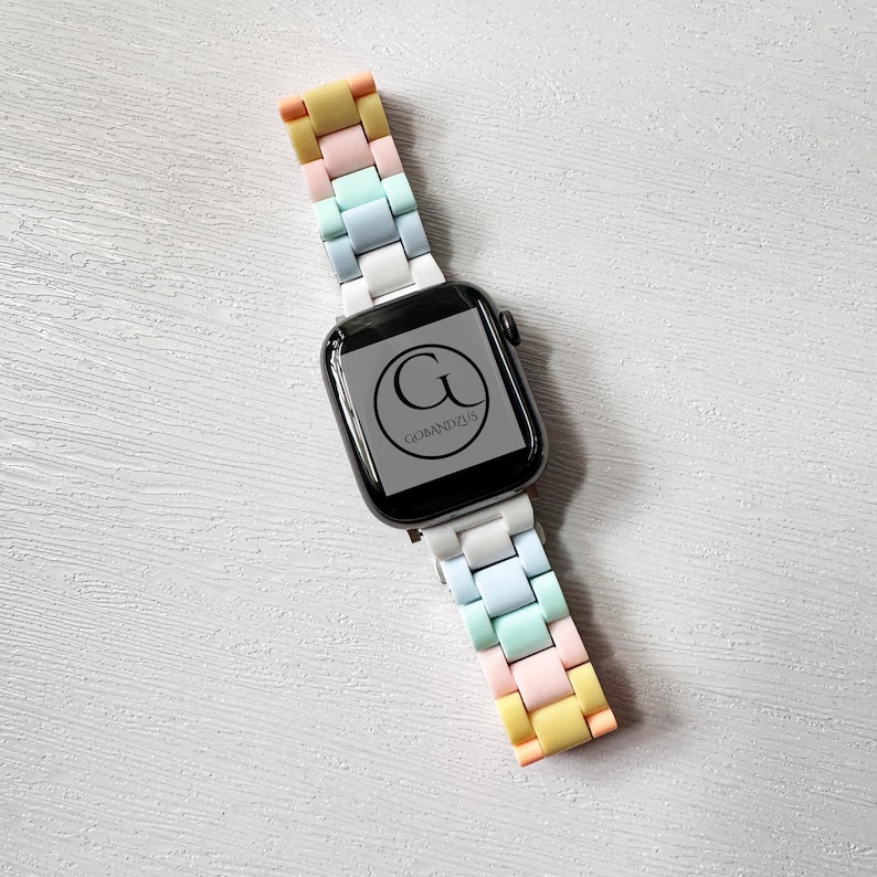 Op de afbeelding: Een witte Apple Watch met een regenboogkleurig siliconen bandje. Het horloge heeft een zwart wijzerplaat met de letter "G" en de tekst "GOMANDZUS" eronder.