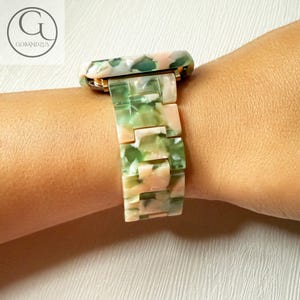 May include: A green and pink patterned watch band with a gold buckle. The band is made of a hard plastic material.