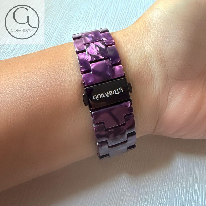 May include: A purple watch band with a marbled pattern and a dark metal clasp. The clasp features the text GOBANDZUS in white lettering. The band consists of rectangular links and is secured around a wrist, showcasing the glossy finish.