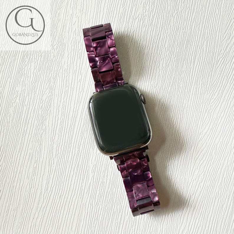 May include: A smartwatch with a dark screen and a link-style watch band made of purple marbled resin. The band features rectangular interlocking pieces with a glossy finish. The logo GOBANDZUS is visible in the upper left corner of the image.