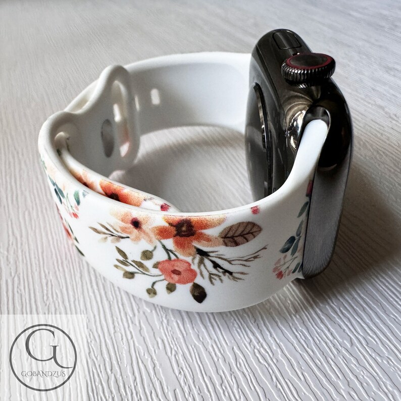 May include: White silicone Apple Watch band with a floral print featuring orange, pink, and green flowers. The band is shown on a black Apple Watch.