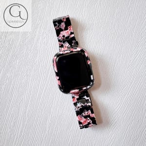 Puede incluir: Un reloj inteligente con pantalla negra y una funda protectora con un patr&oacute;n de resina veteado en negro, rosa y blanco. La correa del reloj consta de eslabones rectangulares de resina a juego con el mismo dise&ntilde;o veteado en negro, rosa y blanco.