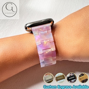 Peut inclure: Un bracelet de montre composé de maillons en résine translucide aux reflets irisés roses et violets avec une finition brillante. Le bracelet est fixé à un boîtier de montre noir. Quatre vignettes circulaires en bas montrent des fermoirs métalliques argentés, dorés, noirs et rose doré avec le texte Custom Engrave Available.