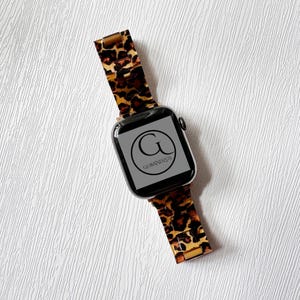 Puede incluir: Un Apple Watch negro con una banda de estampado de leopardo. La esfera del reloj muestra el logotipo "GOBANDZUS" en un c&iacute;rculo.