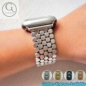 Pode incluir: Uma pulseira de relógio prateada com um design de elos múltiplos e segmentos metálicos arredondados de formato octogonal. A pulseira conecta-se a uma caixa de relógio lisa e curva. Quatro pequenos círculos mostram exemplos de gravação.