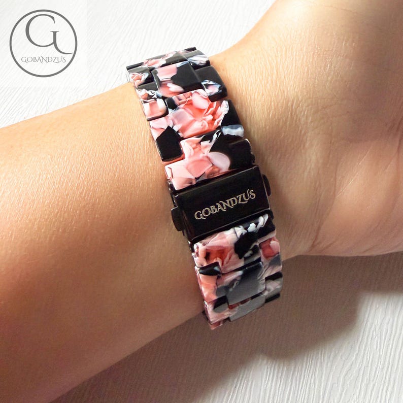 Puede incluir: Una correa de reloj con un patr&oacute;n jaspeado de segmentos negros, blancos y rosa suave. La correa incluye un cierre de metal negro grabado con el texto GOBANDZUS. Los eslabones son rectangulares y pulidos, creando una superficie lisa y continua.