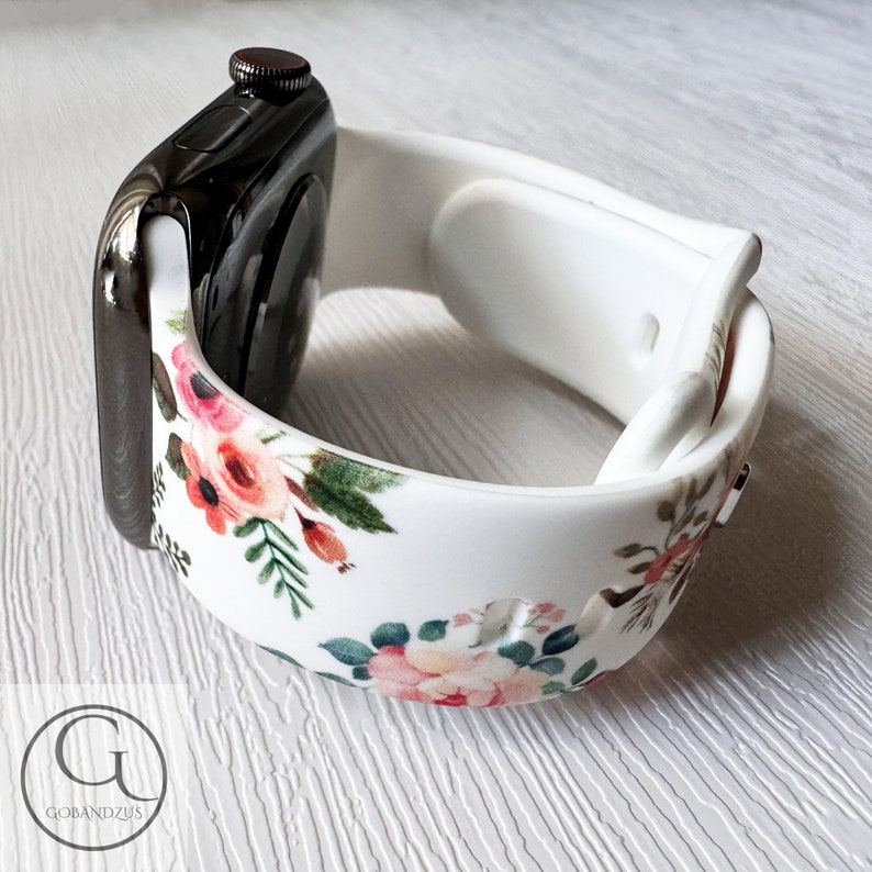 May include: White silicone Apple Watch band with a floral print featuring pink, red, and green flowers. The band is shown on a black Apple Watch.
