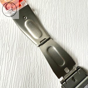 May include: A silver-toned stainless steel watch clasp with a folding mechanism. The metal surface is engraved with the text STAINLESS STEEL. Attached to the clasp is a translucent orange and red watch band segment with a GOBANDZUS logo overlay.
