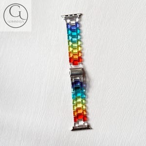 May include: A watch band featuring a series of translucent, rectangular links in a vibrant rainbow gradient of blue, green, yellow, orange, and red. The band includes silver-toned metal connectors at both ends and a central silver-toned clasp.