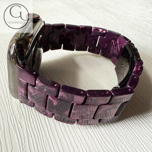 May include: A smartwatch with a dark metallic casing and a link-style watch band featuring a marbled purple and black pattern. The band consists of rectangular segments connected by pins, and the watch includes a black digital display interface.