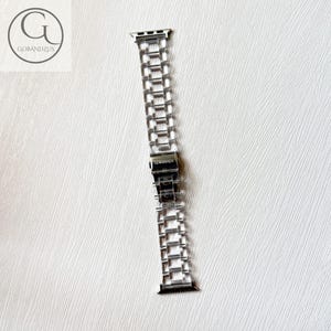 May include: A silver-toned metal watch band featuring a multi-link chain design with a central folding clasp. The clasp is engraved with the text GOBANDZUS. The band includes attachment connectors at both ends for securing to a watch face.