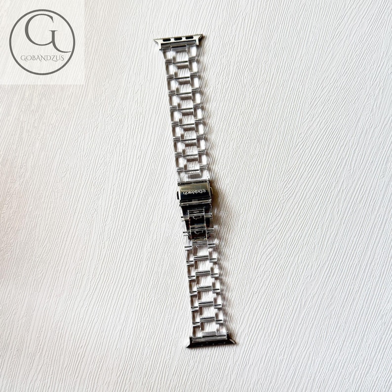 May include: A silver-toned metal watch band featuring a multi-link chain design with a central folding clasp. The clasp is engraved with the text GOBANDZUS. The band includes attachment connectors at both ends for securing to a watch face.