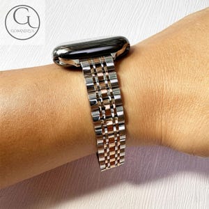 Puede incluir: Una correa de reloj plateada y oro rosa está sujeta a un reloj inteligente negro. La correa está hecha de una serie de eslabones rectangulares. El reloj está en la muñeca de una persona.