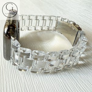 May include: A smartwatch with a dark metallic casing and a clear, transparent acrylic link watch band. The band features a series of rectangular, polished segments connected by metal pins, with a silver-toned metal clasp mechanism visible.
