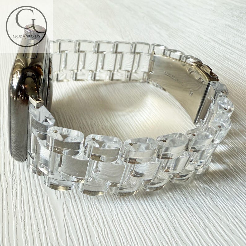 May include: A smartwatch with a dark metallic casing and a clear, transparent acrylic link watch band. The band features a series of rectangular, polished segments connected by metal pins, with a silver-toned metal clasp mechanism visible.