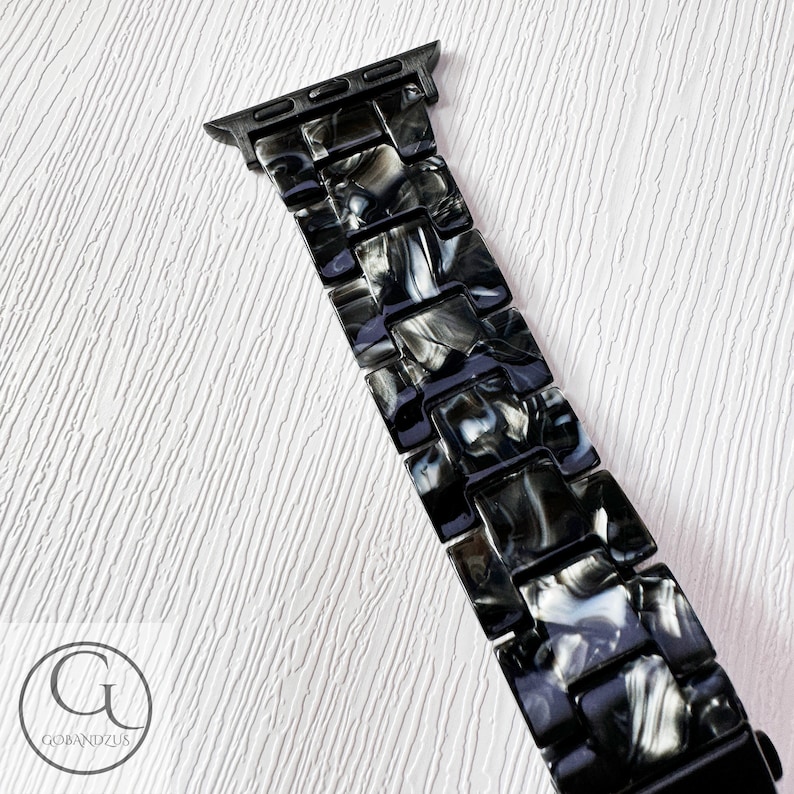 May include: Black and white marbled Apple Watch band with a black metal clasp.