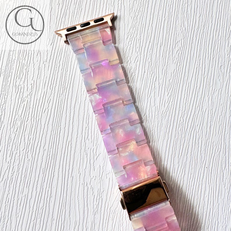 May include: A pink, blue and white patterned Apple Watch band with a rose gold metal connector.