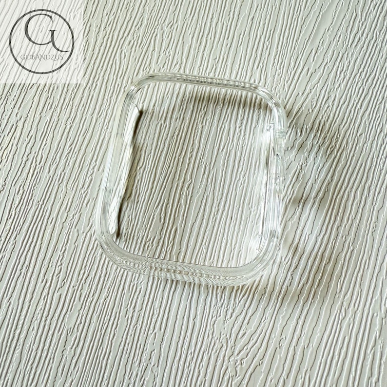 May include: A clear, rectangular protective watch case with rounded corners and a hollow center. The frame is transparent and smooth, designed to fit over the face of a digital watch. The logo GOLANDZUS is visible in the upper left corner.