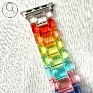May include: A watch band featuring translucent, multi-colored resin links in a gradient of red, orange, yellow, green, blue, and purple. The band includes a silver-toned metal connector piece at the top for attachment to a digital watch device.