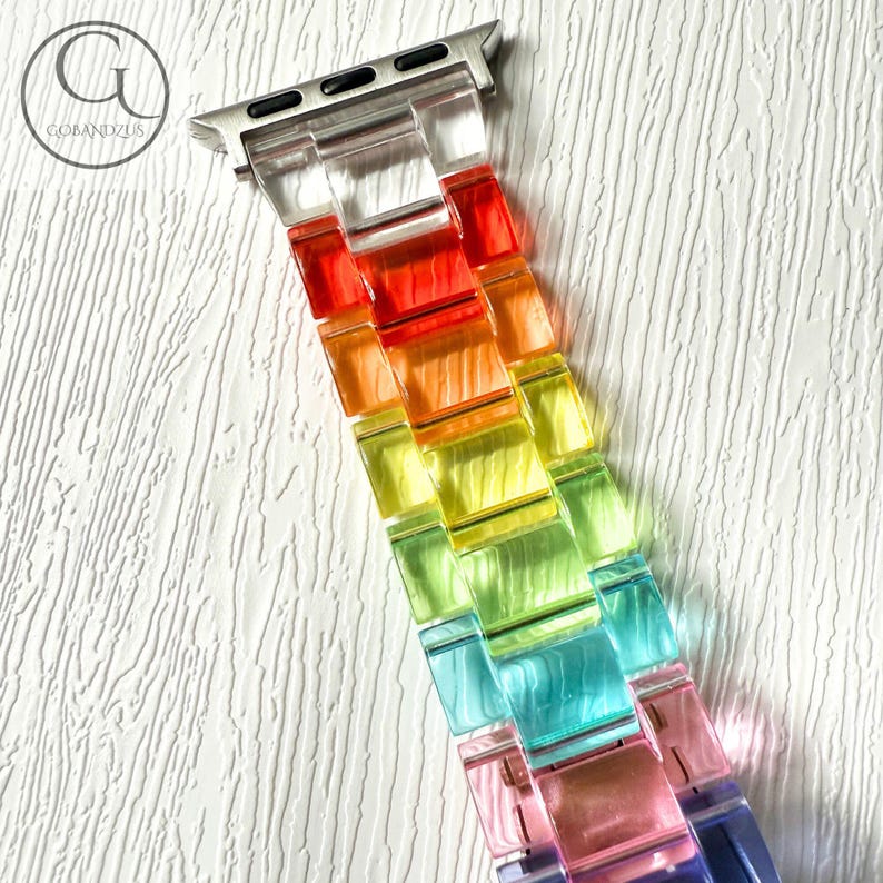 May include: A watch band featuring translucent, multi-colored resin links in a gradient of red, orange, yellow, green, blue, and purple. The band includes a silver-toned metal connector piece at the top for attachment to a digital watch device.