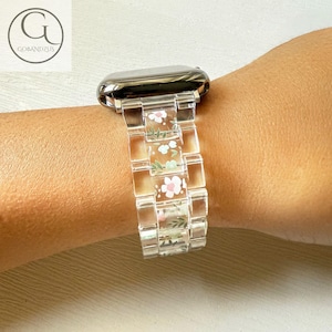 Könnte beinhalten: Transparenter Uhrenarmband mit floralem Design. Das Armband besteht aus transparentem Kunststoff und weist kleine rosa und weiße Blumen mit grünen Blättern auf. Die Uhr hat ein silberfarbenes Gehäuse.