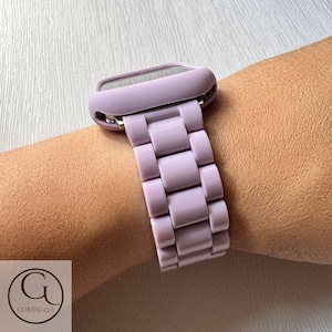Könnte beinhalten: Ein helllila Uhrenarmband und ein Uhrengehäuse sind an einem Handgelenk zu sehen. Das Uhrenarmband besteht aus rechteckigen Gliedern. Das Uhrengehäuse ist quadratisch mit abgerundeten Kanten.