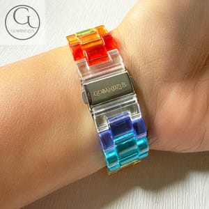 May include: A rainbow-colored watch band made of translucent links in red, orange, yellow, green, blue, and purple. The band features a metallic silver-toned clasp engraved with the text GOBANDZUS, securing the links together on the wrist.