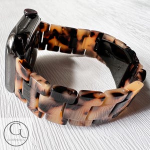 May include: A brown and black tortoiseshell patterned watch band with a black metal clasp. The band is made of a hard plastic material and has a glossy finish.